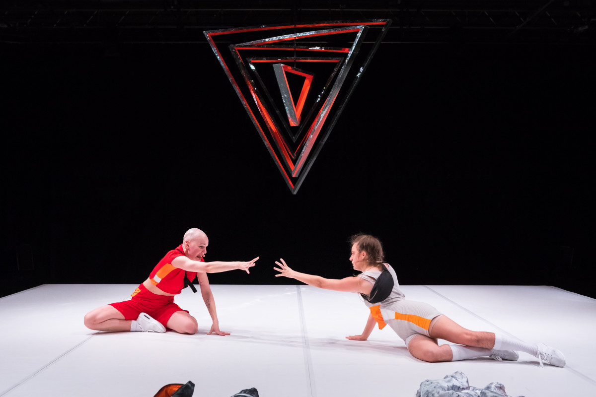 Auf dem Foto sitzen die beiden Tänzer:innen Lisa Rykena und Carolin Jüngst mittig auf einer weißen Fläche vor schwarzem Hintergrund. Hinter ihnen schwebt ein Objekt in Dreiecksform, welches silbern und rot schimmert und spitz nach unten zeigt. Lisa hat eine Glatze und weiße blasse Hautfarbe. Sie trägt ein rotes, eng anliegendes Oberteil mit orangenen, dicken Streifen an der Seite und eine rote, kurze enge Hose. Carolin sitzt rechts von Lisa. Sie hat weiße Hautfarbe, Ihre braunen Haare sind zu einem Zopf gebunden. Sie trägt als Kostüm einen arm- und  beinfreien Einteiler in grau mit orangenen Streifen an der Seite. Lisa und Carolin tragen beide schwarze Mikrofontaschen um die Brust, Headset Mikrofone am Kopf und weiße Socken mit weißen Turnschuhen. Beide Tänzer*innen sind im Sitzen einander zugewandt und versuchen mit einem weit ausgestrecktem Arm einander zu erreichen und zu greifen. Das Gesicht von Lisa ist wie zu einem Weinen verzerrt und der Mund geöffnet.